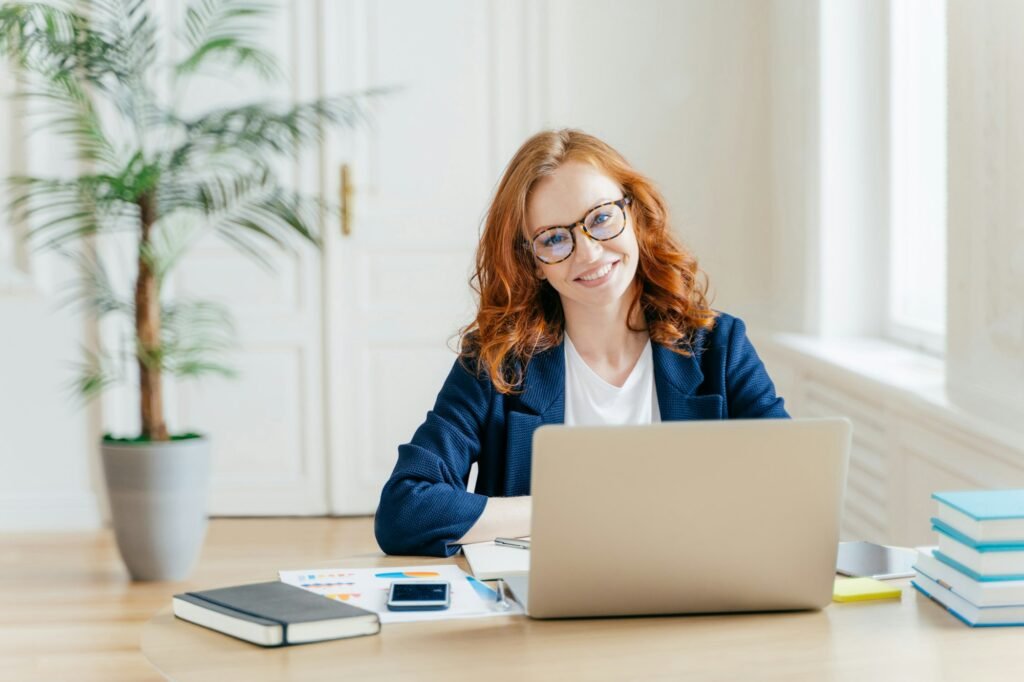 Businesswoman wears glasses for vision correction, sits at desktop in front of opened laptop